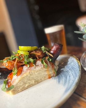 Hot dog, topped with sticky prawns, chilli sauce, coriander. in a bun. Served on a plate with a half pint of beer behind.